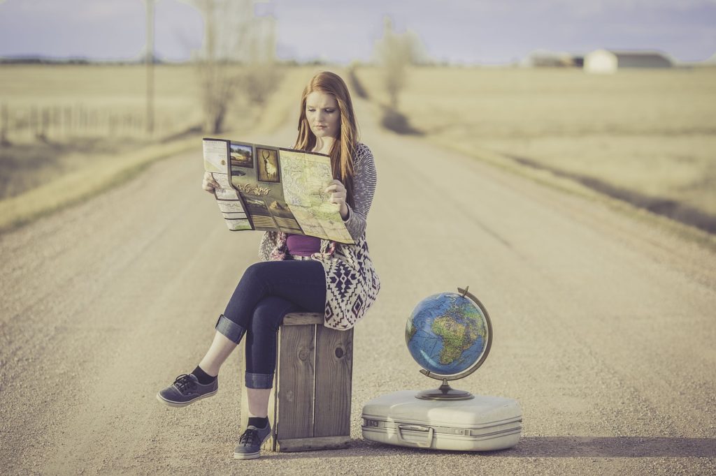 Woman in the middle of a dirt road sitting on a suitcase reading a map next to a globe