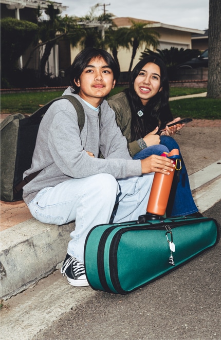 Two teens sitting on a curb next to a green violin case