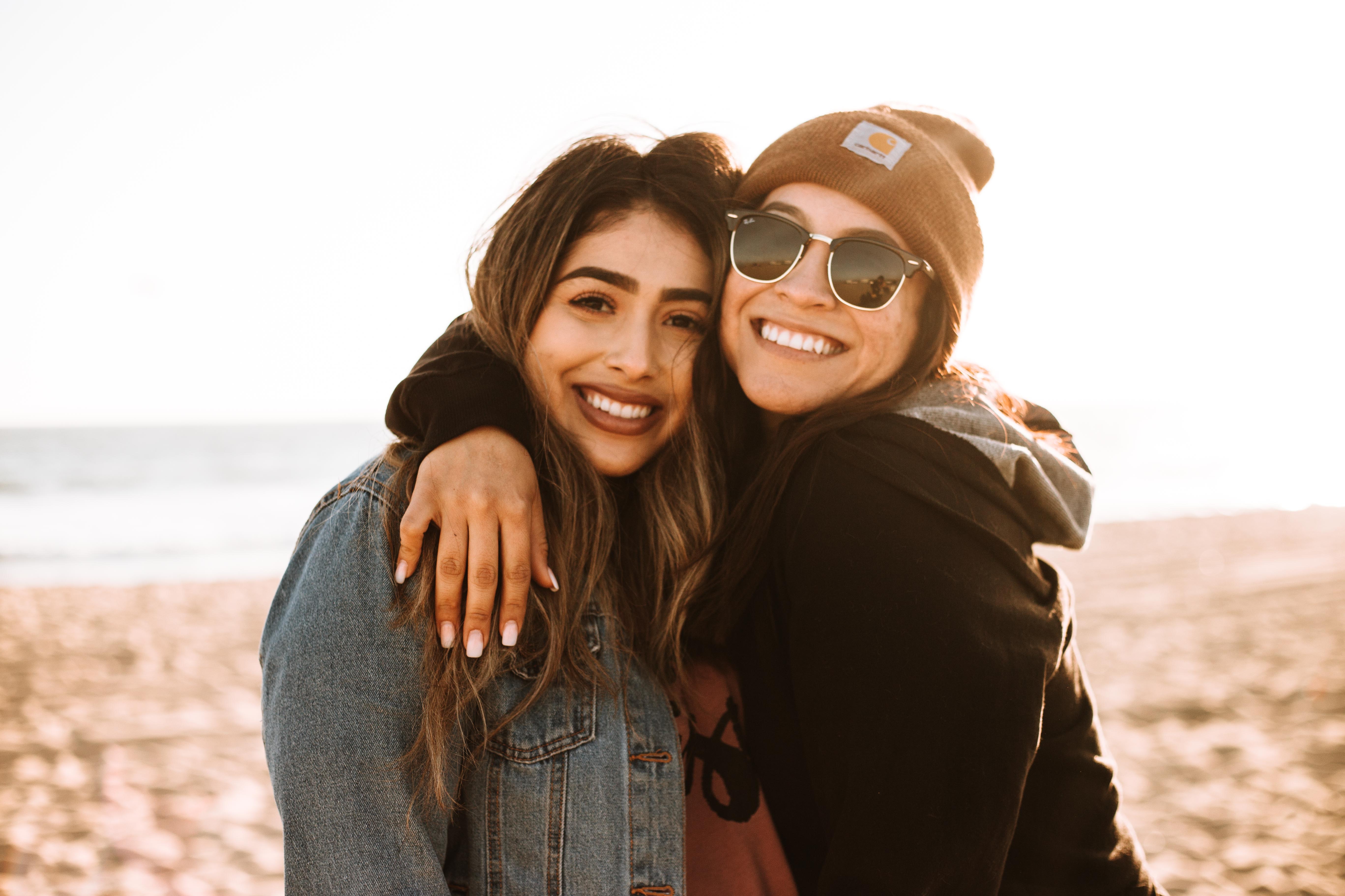 Two women smiling at the beach with their arms around each other.