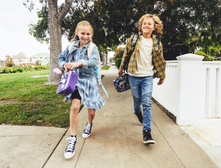 Two children with backpacks skipping on a suburban sidewalk.